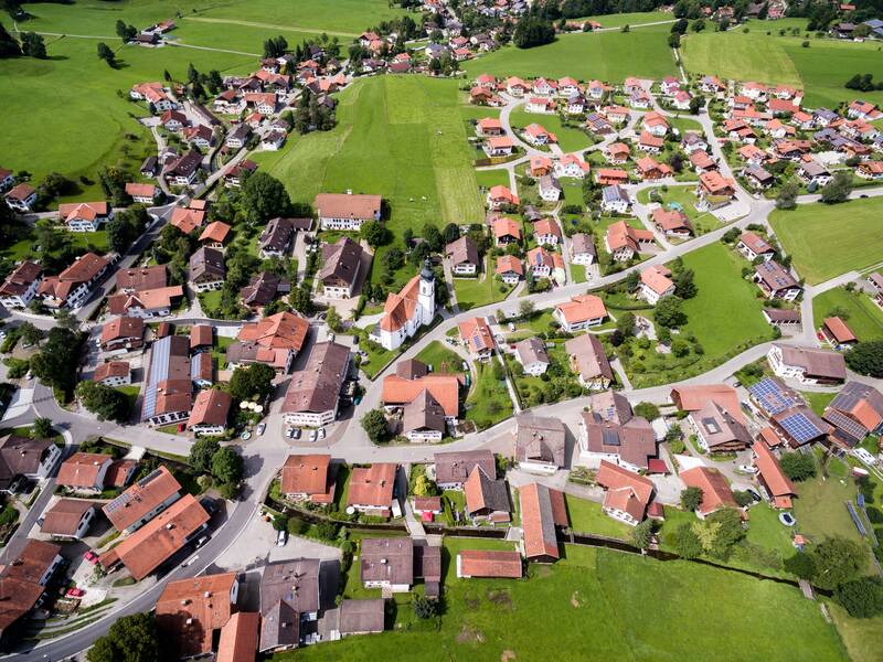 Kommune von oben fotografiert. Hausdächer von Wohnhäusern und die Straßen dazwischen. Um die Häuser herum sind viele, große, grüne Wiesen. 