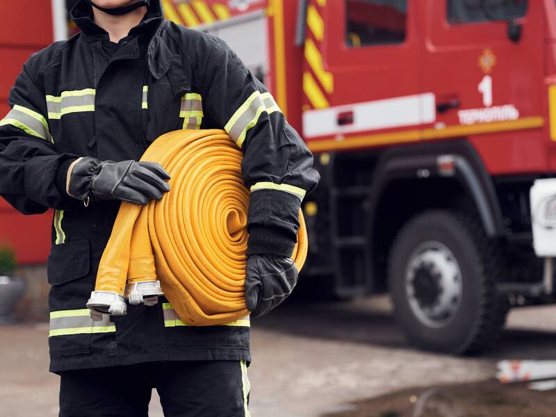 Im Vordergrund steht ein Feuerwehrmann in seiner Ausrüstung und er hat einen aufgerollten Feuerwehrschlauch unter dem Arm. Im Hintergrund steht ein großes Feuerwehrauto. 