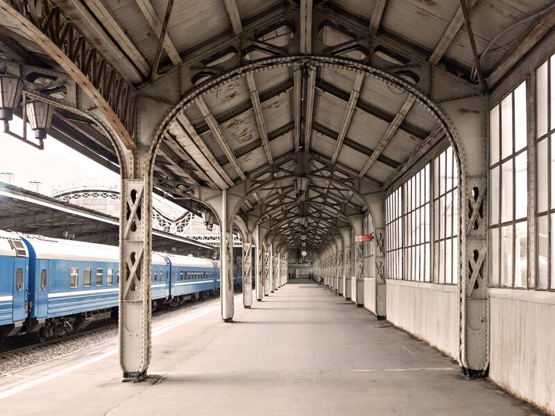 Ein leerer Bahnsteig der überdacht ist. das Dach hat ein aus Stahl bestehendes Fundament welches rund über den Bahnsteig geht. Links auf den gleisen steht ein alter Zug in blau. 
