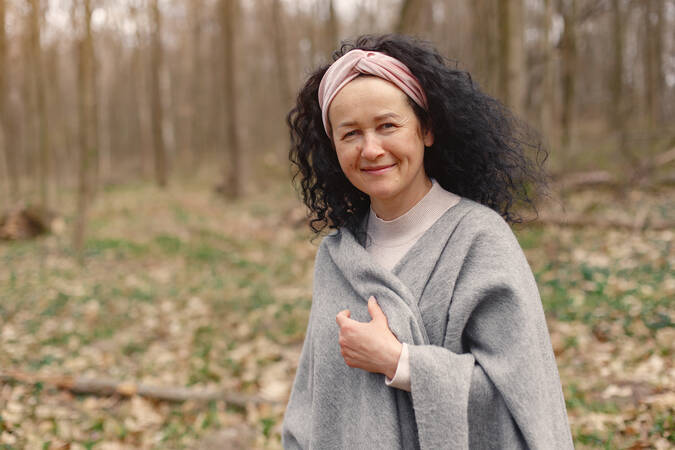 Eine Frau mittleren Alters mit lockigen dunklen Haaren und einem rosa Stirnband in den Haaren. Sie hat graues Oberteil an und hält dieses mit einer Hand vorne an der Brust zusammen. Sie schaut lächeln in die Kamera. Im Hintergrund ist unscharf zu erkennen, dass Sie sich in einem Laubwald aufhält.