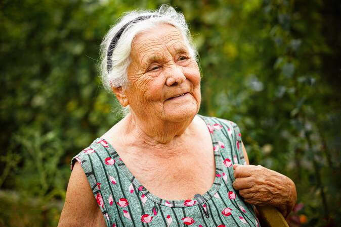 Alte Frau die vor einem Busch steht. Sie hat weiße Haare und darin ein schwarzes Band. Sie trägt ein grünes Top mit roten Blumen darauf. Ihre eine Hand hält sie zu ihrer Schulter hoch. Sie schaut an der Kamera vorbei und lächelt etwas. 