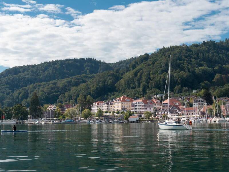 Ein See mit dunklem Wasser im Vordergrund. Auf dem Wasser ist ein stand up paddeling-Brett und ein Mensch. Weiter rechts und weiter nach hinten sind mehrere Segelboote. Im Hintergrund sind Wohnhäuser