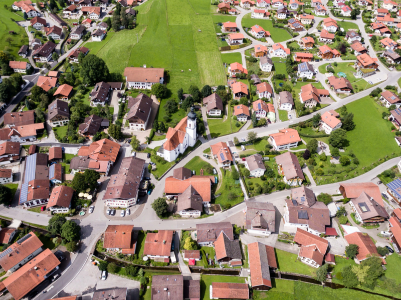 Eine Gemeinde von oben. Es sind mehrere Häuser mit roten Dächern und Straßen. Zwischendrin sind große grüne Wiesen-Felder. 
