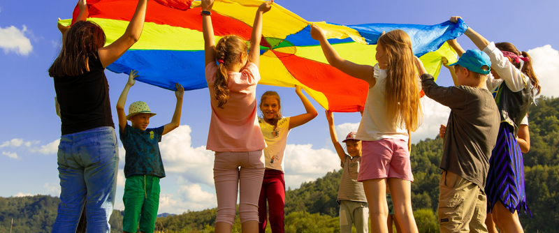 Mehrere Kinder stehen in einem kreis und halten ihre Hände nach oben. Sie halten alle zusammen ein großes, rundes, buntes Tuch über ihre Köpfe. Sie schauen sich dabei unter dem Tuch durch an. 