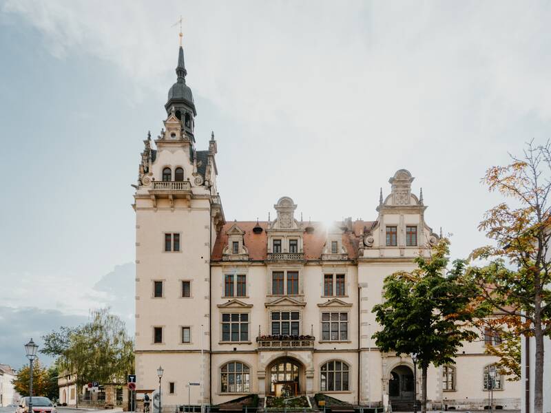 Kirche von vorne. Auf der linken Seite des Gebäudes ist der große Kirchturm. Vorne ist der große Eingang mit einer Treppe. Rechts im Vordergrund stehen Bäume. 