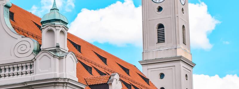 Eine Kirche die zur Hälfte von einer schrägen Perspektive abgebildet ist. Weiter hinten sieht mit der komplette Kirchturm auf dem Bild. Im Vordergrund ist ein Biergarten mit Schirmen und einem Schild mit der Restaurantbezeichnung "Cafe Kaiserschmarrn".
