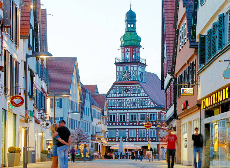 Fußgängerzone einer beleuchteten Stadt. Geradeaus ist die Kirche und rechts und links Ladengeschäfte. Es sind vereinzelt Menschen in der Fußgängerzone unterwegs. 