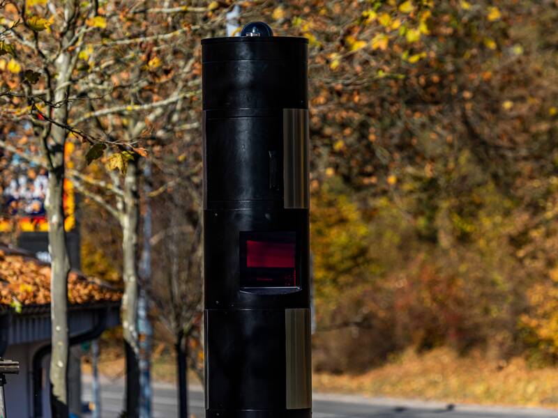 Eine schwarze Blitzersäule. Im Hintergrund sind herbstliche Bäume und Sträucher.