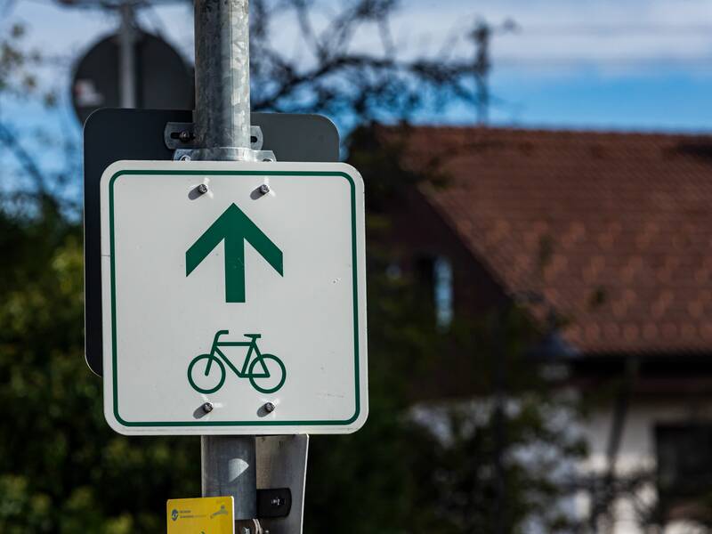 Ein weißes Schild mit einem grünen Fahrrad und Richtungspfeil als Wegweiser für einen Radweg.  Das Schild ist an einem Laternenpfosten befestigt.