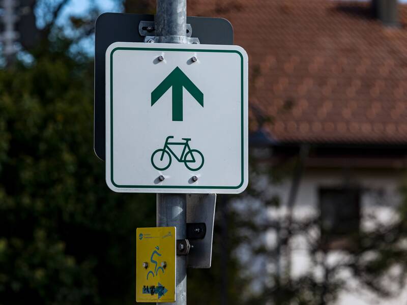 Ein weißes Schild mit einem grünen Fahrrad und Richtungspfeil als Wegweiser für einen Radweg. Das Schild ist an einem Laternenpfosten befestigt.