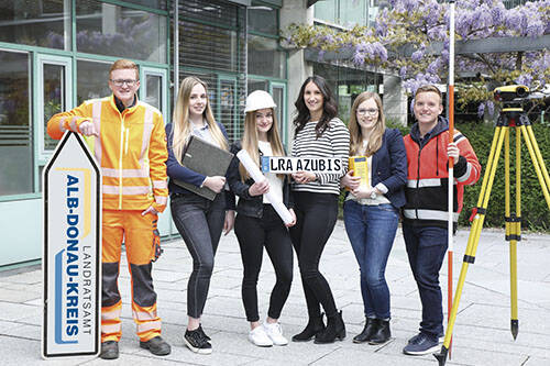 Eine Gruppe von Menschen steht vor einem Bürogebäude, von denen einige Arbeitskleidung tragen, während eine Person ein Schild mit der Aufschrift "LRA Azubis" hält.