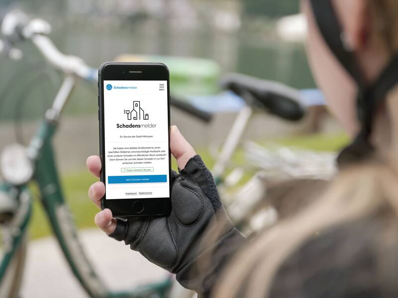 Eine Person hält ein Smartphone, das die Webseite "Schadensmelder" anzeigt, während im Hintergrund ein Fahrrad unscharf zu sehen ist.