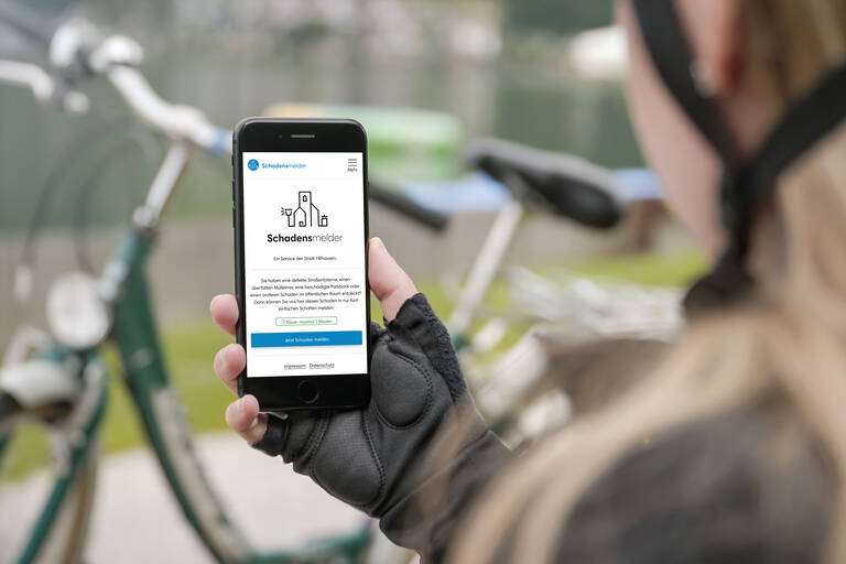 Eine Person hält ein Smartphone, das die Webseite "Schadensmelder" anzeigt, während im Hintergrund ein Fahrrad unscharf zu sehen ist.